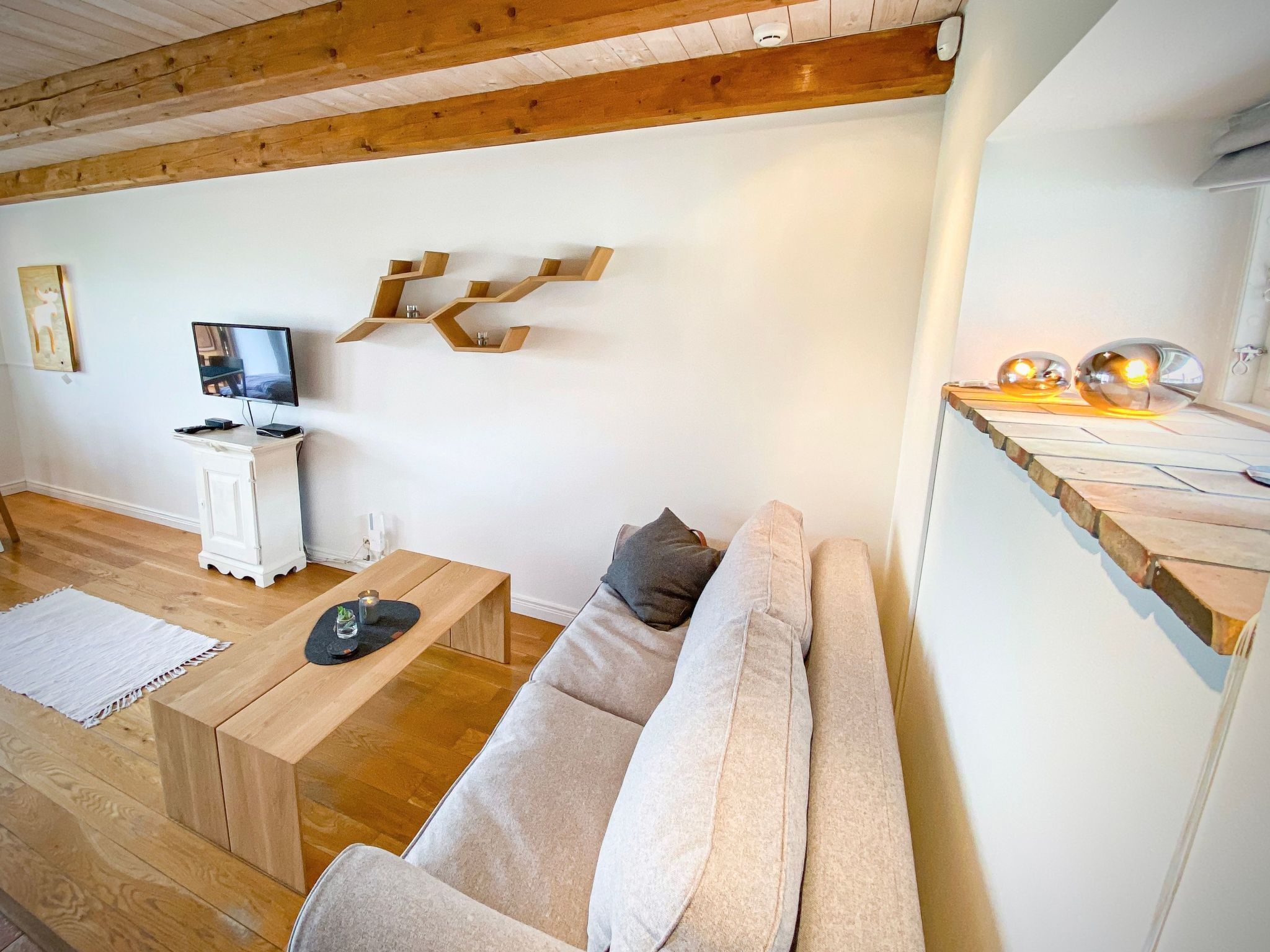 A modern living room with a wooden ceiling, a white wall, a wooden coffee table, a beige sofa, a TV on a white cabinet, and a unique wooden shelf with spherical lights.