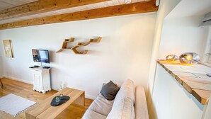 A modern living room with a wooden ceiling, a white wall, a wooden coffee table, a beige sofa, a TV on a white cabinet, and a unique wooden shelf with spherical lights.
