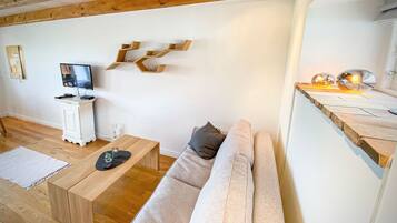 A modern living room with a wooden ceiling, a white wall, a wooden coffee table, a beige sofa, a TV on a white cabinet, and a unique wooden shelf with spherical lights.