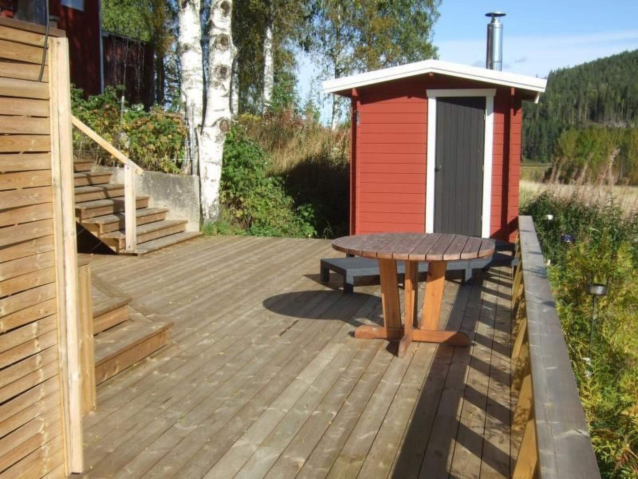 A wooden deck with a table and chairs, a red building with a door, and a staircase leading up.