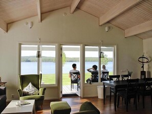 Ruime kamer met een grote schuifpui die uitkomt op een terras met uitzicht op een meer. Binnenin staat een groene fauteuil, een eettafel en een bijzettafeltje.