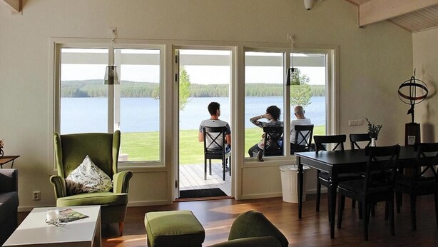 Il s’agit d’une pièce spacieuse dotée d’une grande porte-fenêtre coulissante qui donne sur une terrasse offrant une vue sur un lac. On y trouve un fauteuil vert, une table à manger et une table basse.
