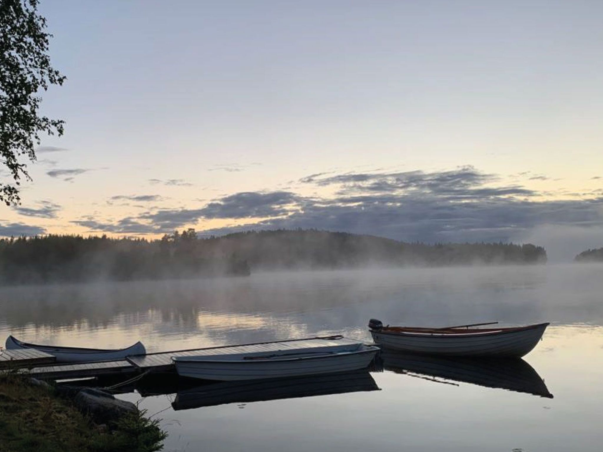 A calm lake with boats docked on the shore and a forested hill in the background.