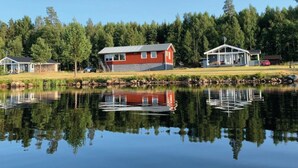 En stuga vid sjön med en röd fasad, en brygga och en spegelbild i vattnet.