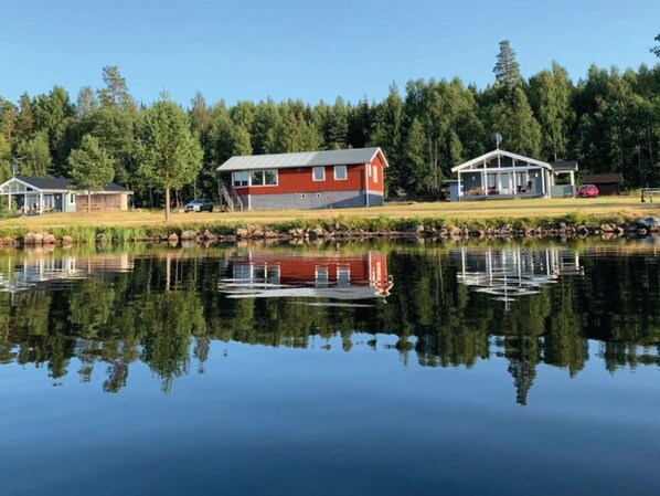 Una cabaña junto al lago con fachada roja, un muelle y su reflejo en el agua.