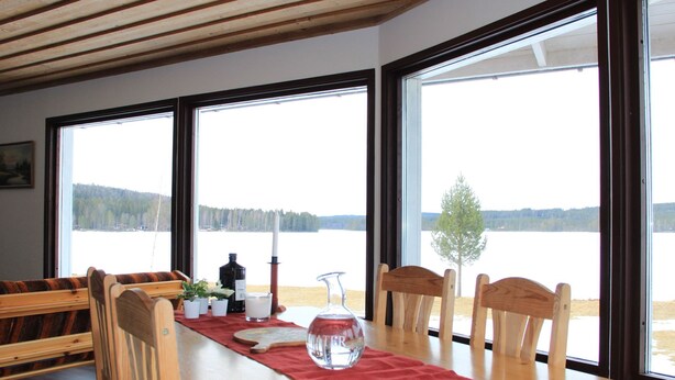 En matplats med ett träbord, stolar och utsikt över en sjö genom stora fönster.