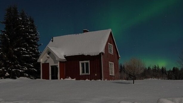 Una casa de madera roja con tejado cubierto de nieve, rodeada de nieve y árboles, bajo un cielo nocturno con aurora boreal verde.