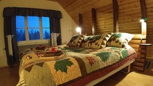 Un dormitorio acogedor con una cama grande, vigas de madera en el techo, una ventana con cortinas y una mesita de noche con lámpara.