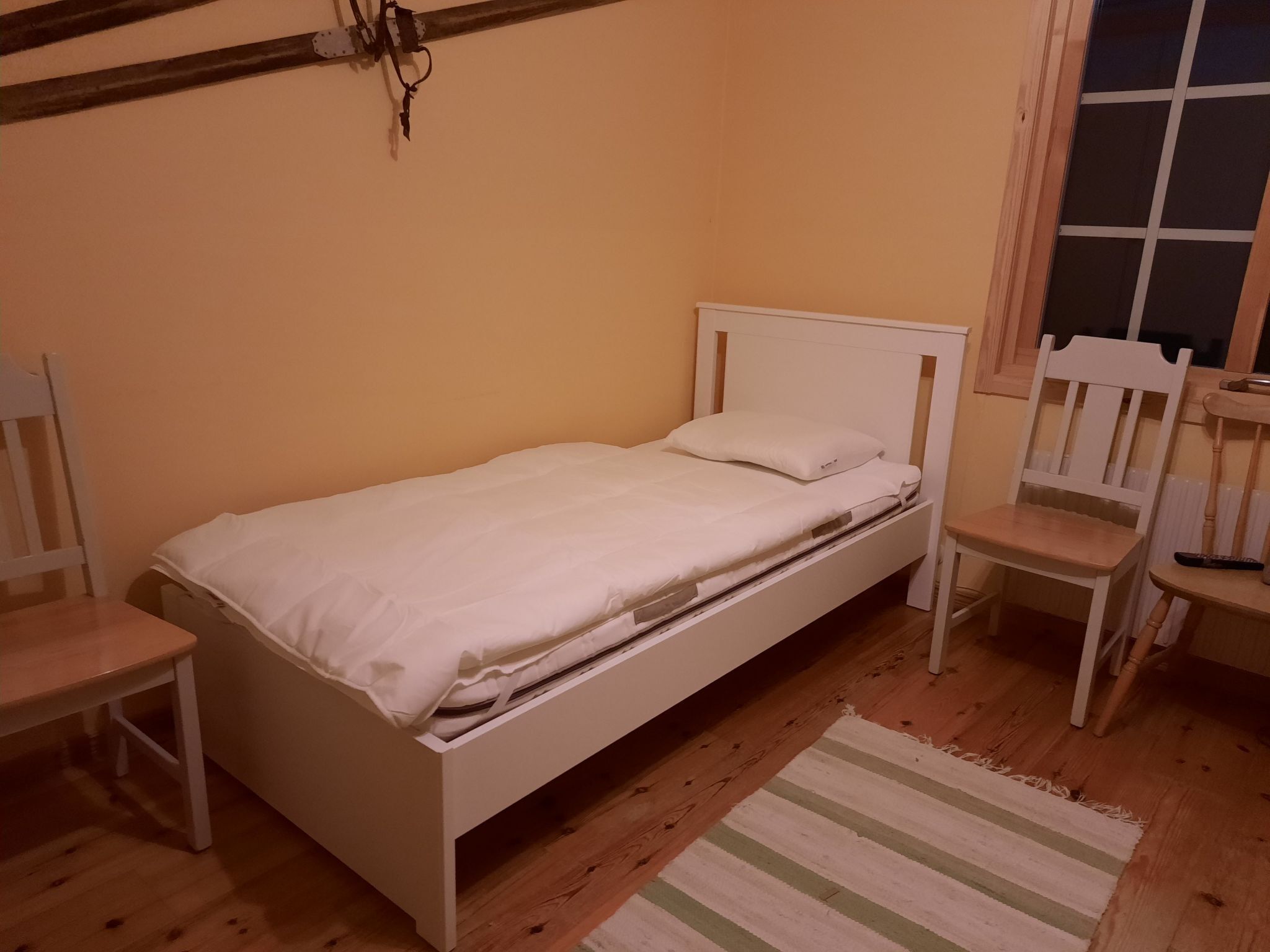 A small, single-bed room with a wooden floor, a white bed, a wooden chair, and a window with blinds.