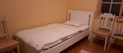A small, single-bed room with a wooden floor, a white bed, a wooden chair, and a window with blinds.