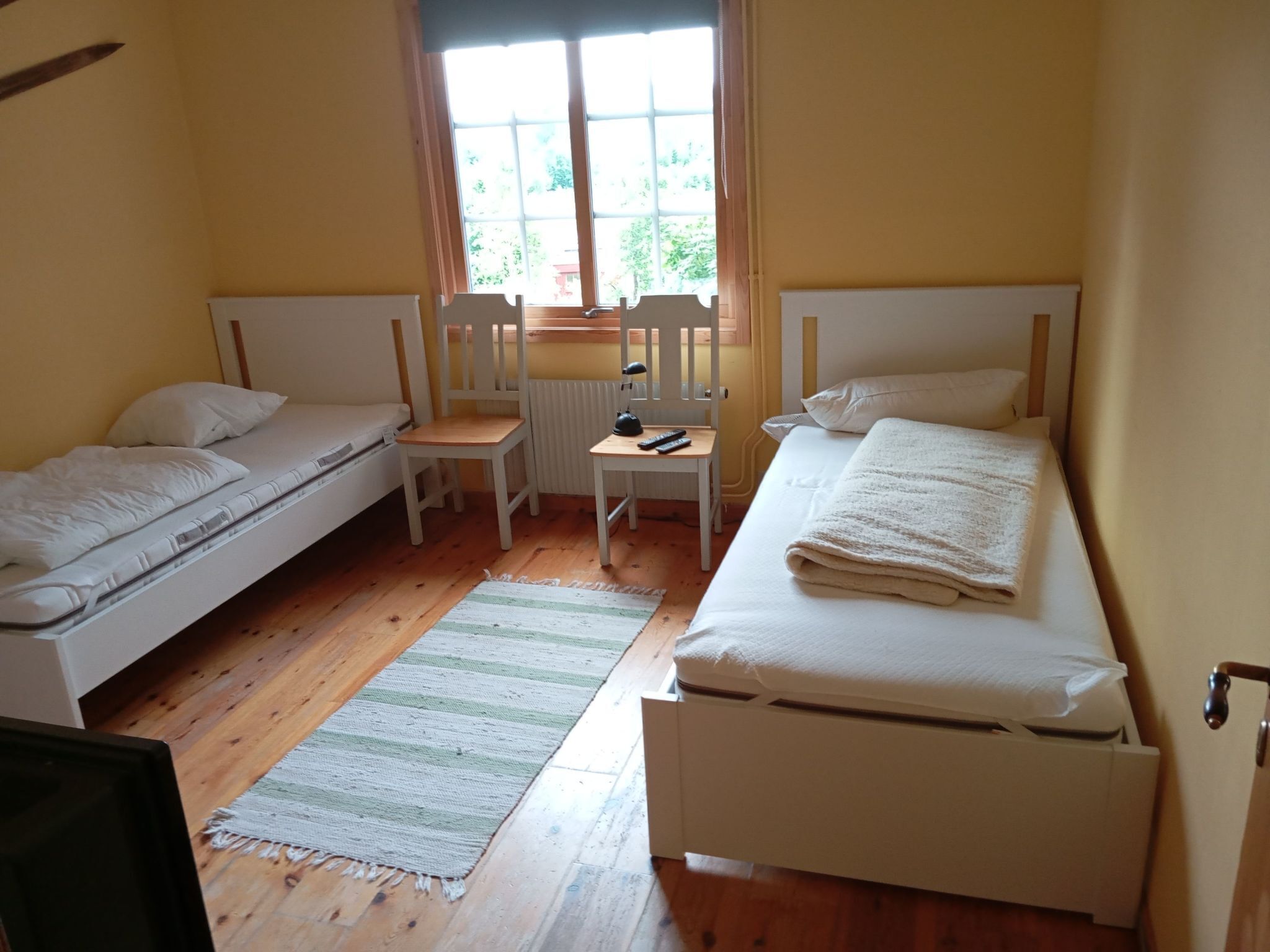 Une chambre avec deux lits simples, une petite table avec une chaise, un radiateur et une fenêtre avec des rideaux.