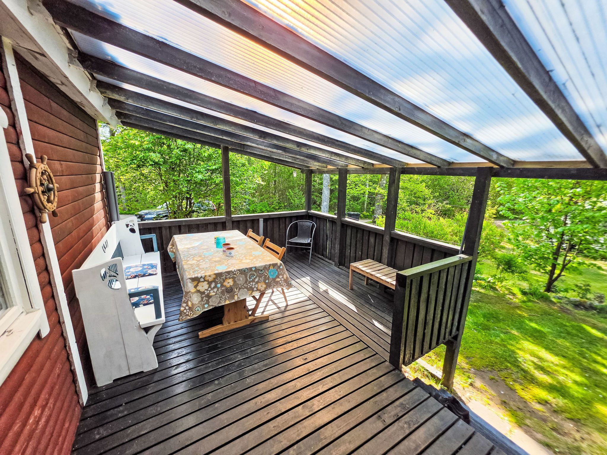 A covered wooden deck with a table and chairs, surrounded by greenery.