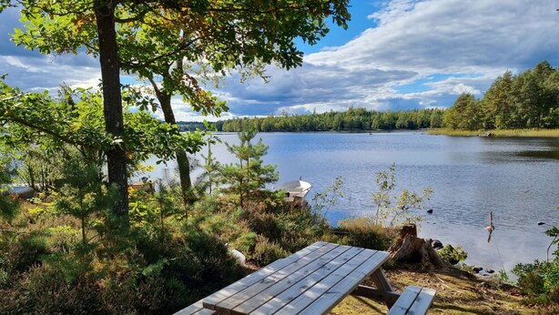 A wooden picnic table by a lake with trees and a boat in the background. - Gemütliches Ferienhaus in Seenähe by Interhome (Hallaryd)
