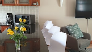 A modern kitchen with a black countertop, a coffee maker, and a microwave. There is a dining table with white chairs and a vase with yellow flowers. A grey sofa with patterned cushions is in the living area. - in Kirstenhof mit Privatem Garten by Interhome (Kapstadt)