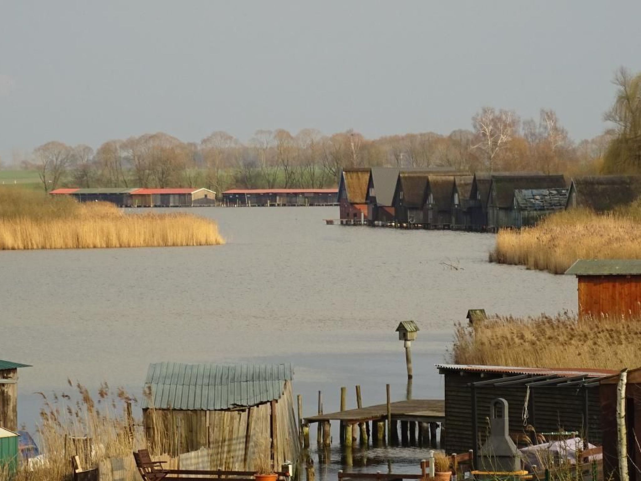 Zona ribereña con construcciones de madera, que incluyen un muelle y pequeños edificios, rodeada de hierbas altas.