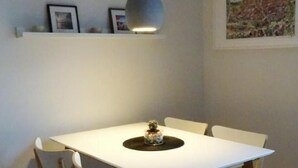 En matplats med ett vitt bord, fyra stolar, en hänglampa och en hylla med inramade bilder.