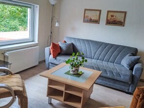 Un salon avec un canapé gris, une table basse en bois sur laquelle est posée une plante, un radiateur, une fenêtre et deux tableaux encadrés accrochés au mur.