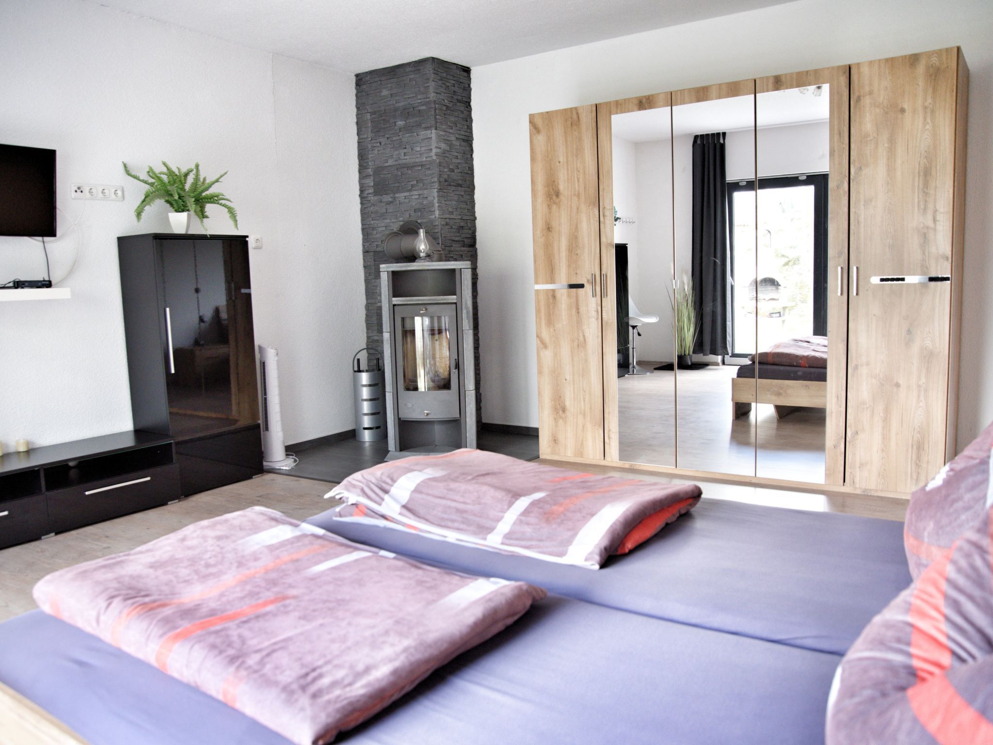 Ein modernes Schlafzimmer mit großem Kleiderschrank, Fernseher, Pflanze und einem Bett mit Kissen.
