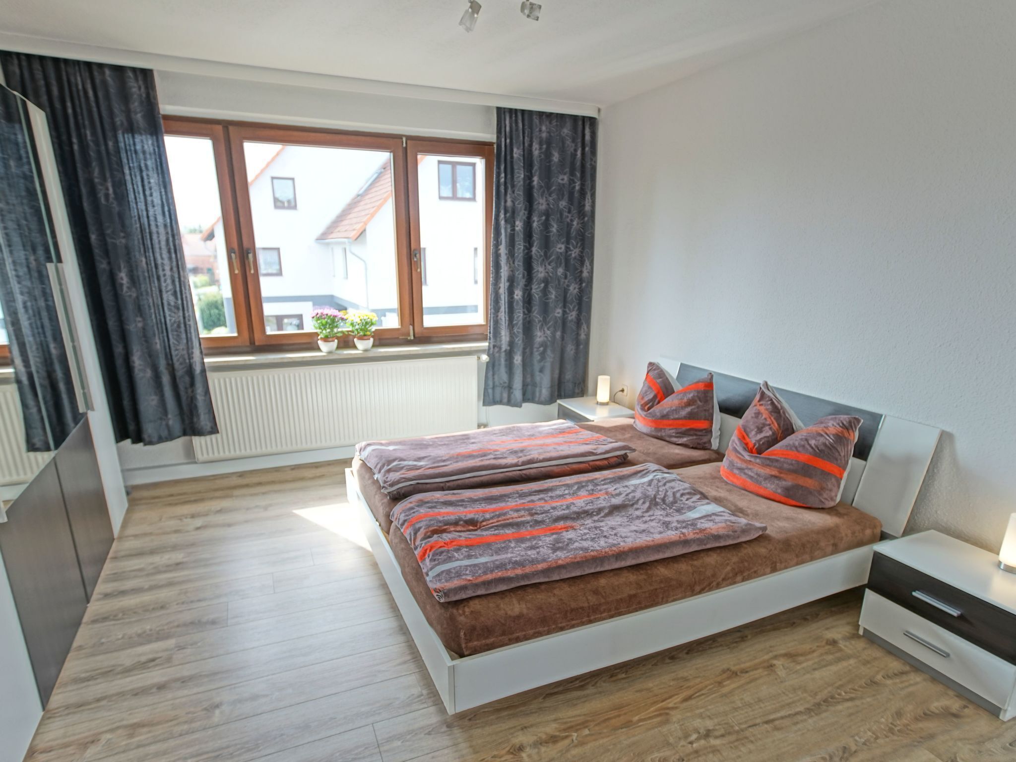 A modern bedroom with a large bed, wooden flooring, a window with curtains, a bedside table, and a nightstand.