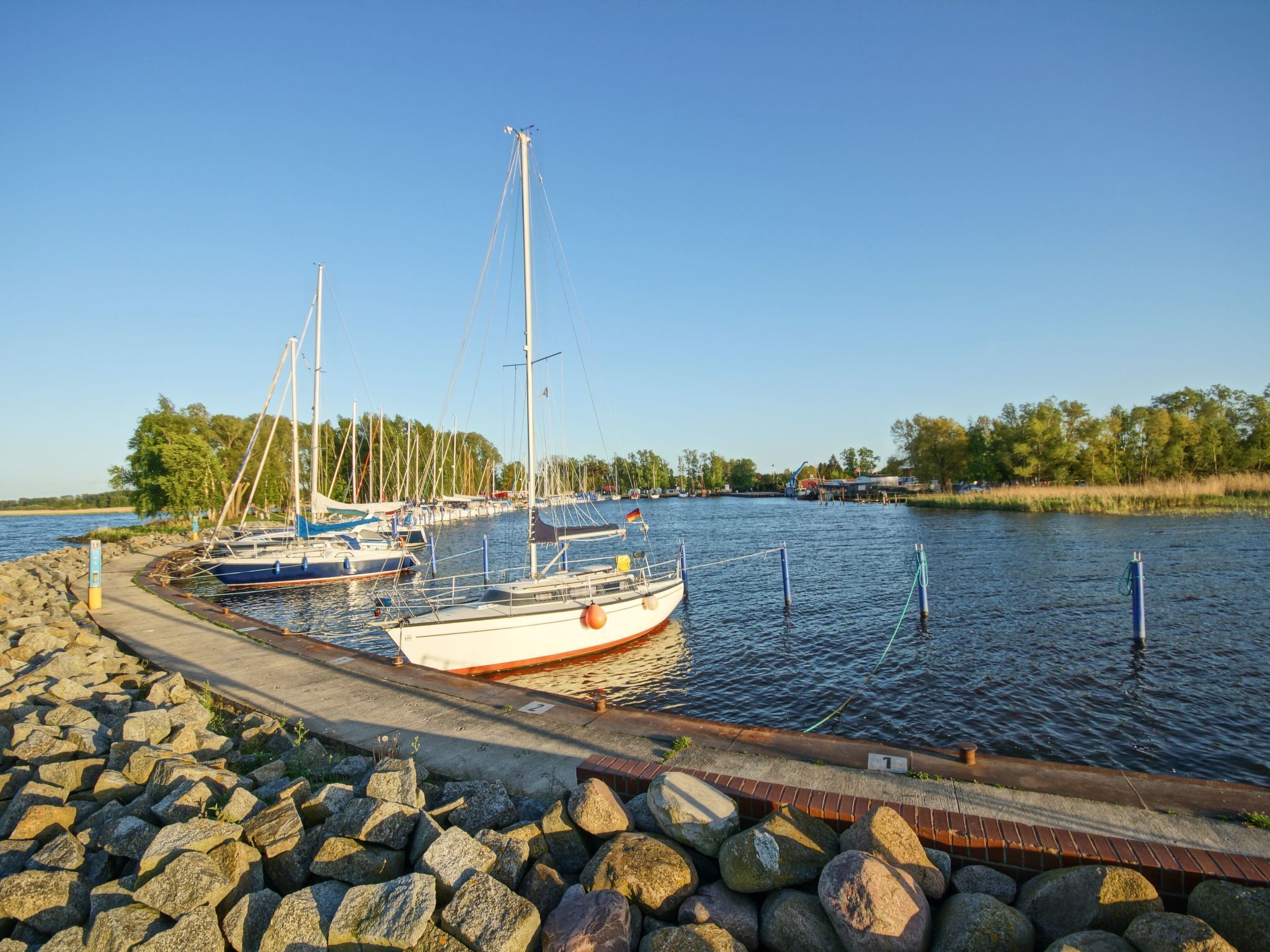 A marina with several sailboats docked, a concrete walkway, and a rocky shoreline.