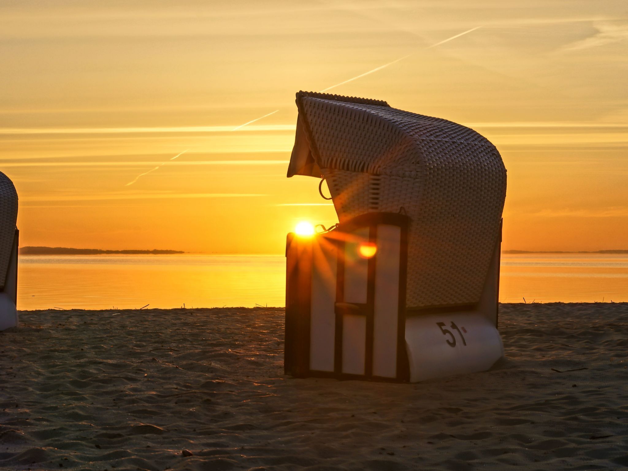 Una silla de playa con el número "51" en el lateral, ubicada en una playa arenosa con un atardecer de fondo.