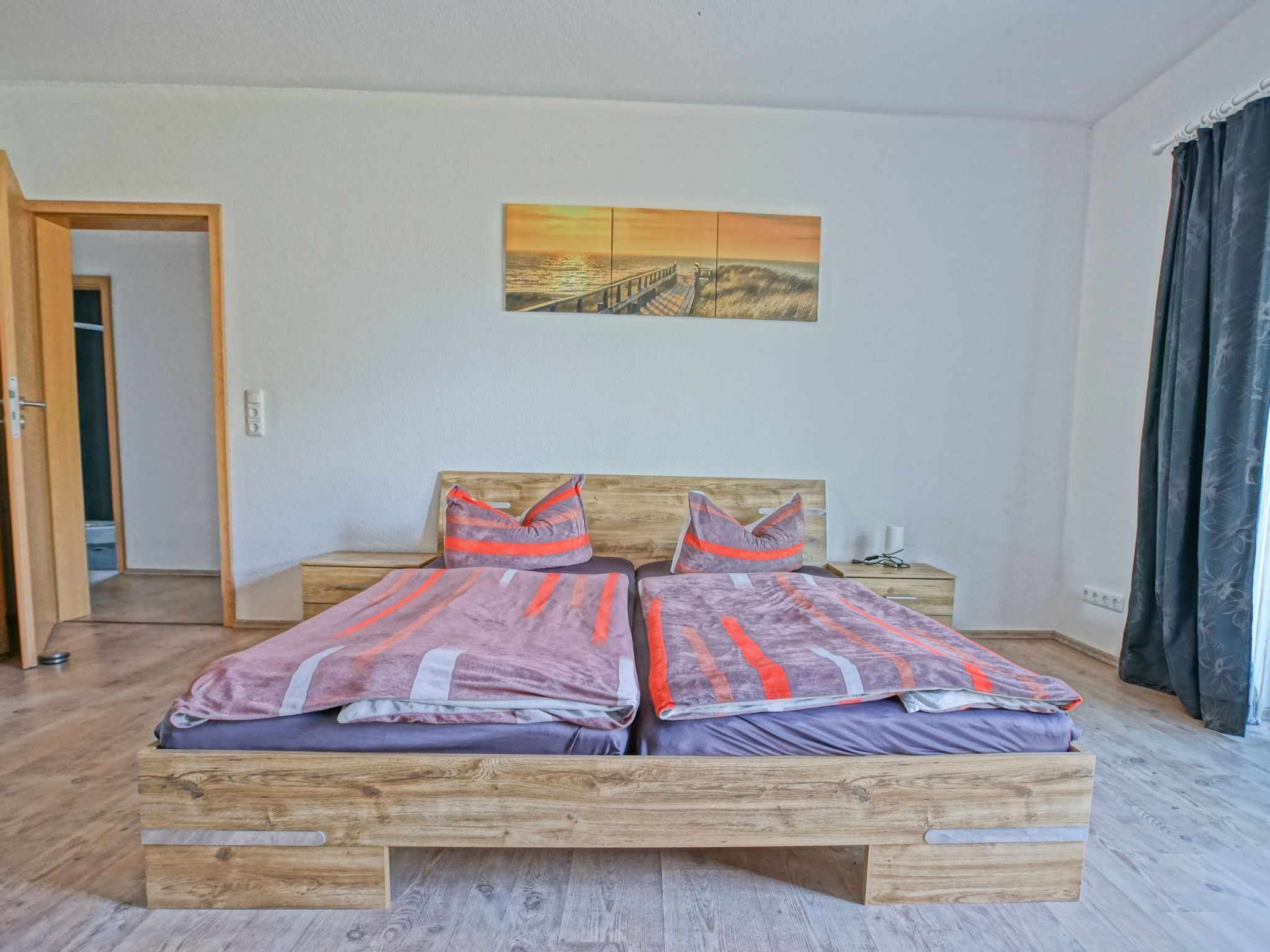 Un lit double avec un cadre en bois, deux oreillers et une couverture à rayures orange. Un tableau représentant un coucher de soleil est accroché au mur au-dessus du lit.