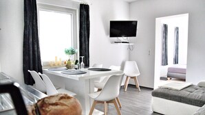 Een moderne, goed verlichte kamer met een eethoek, een flatscreen televisie aan de muur en een gezellige zithoek met een bank en een fauteuil.