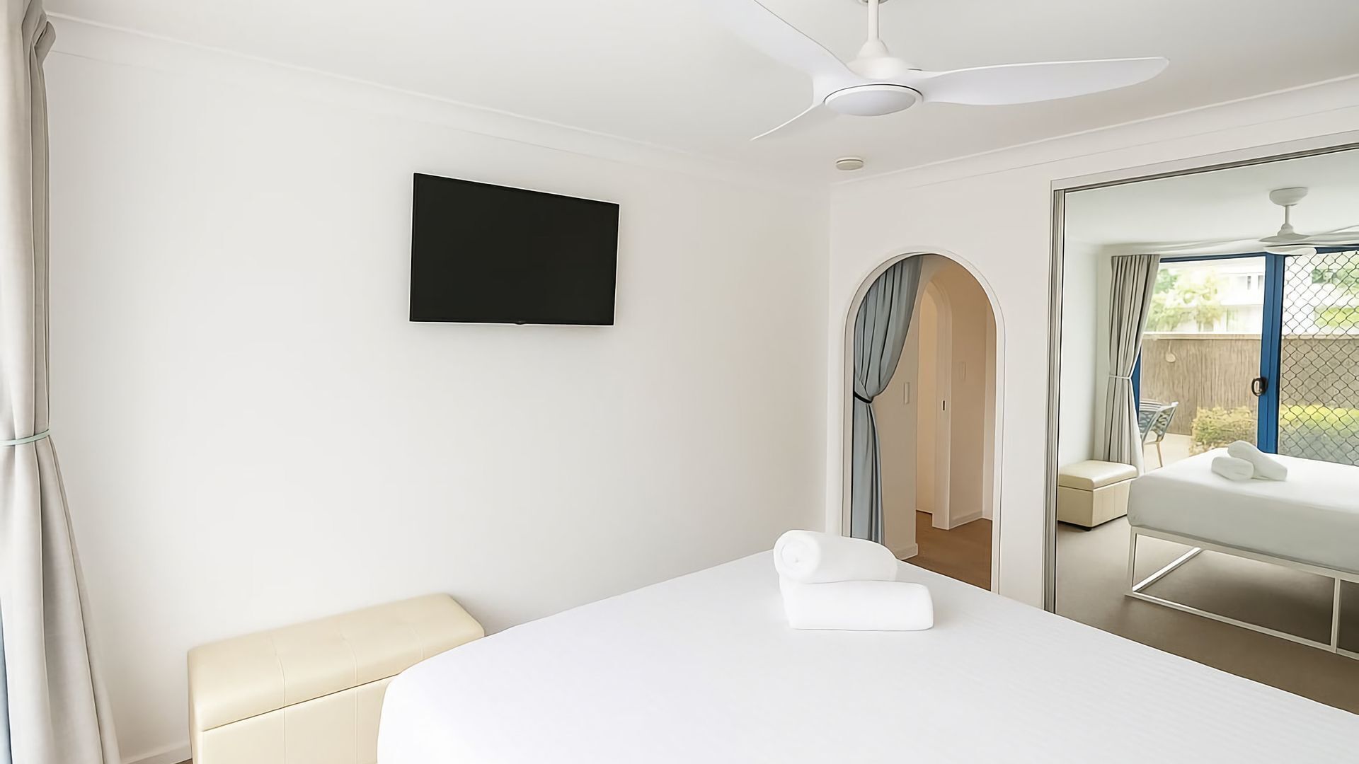 Et moderne hotelværelse med seng, fladskærms-tv, loftventilator og udsigt gennem en buet døråbning.
