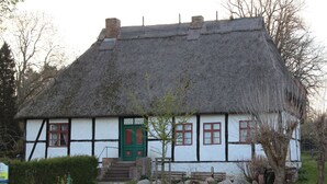 Ein traditionelles Haus mit Strohdach, einer grünen Tür, weißen Wänden und einer kleinen Veranda.