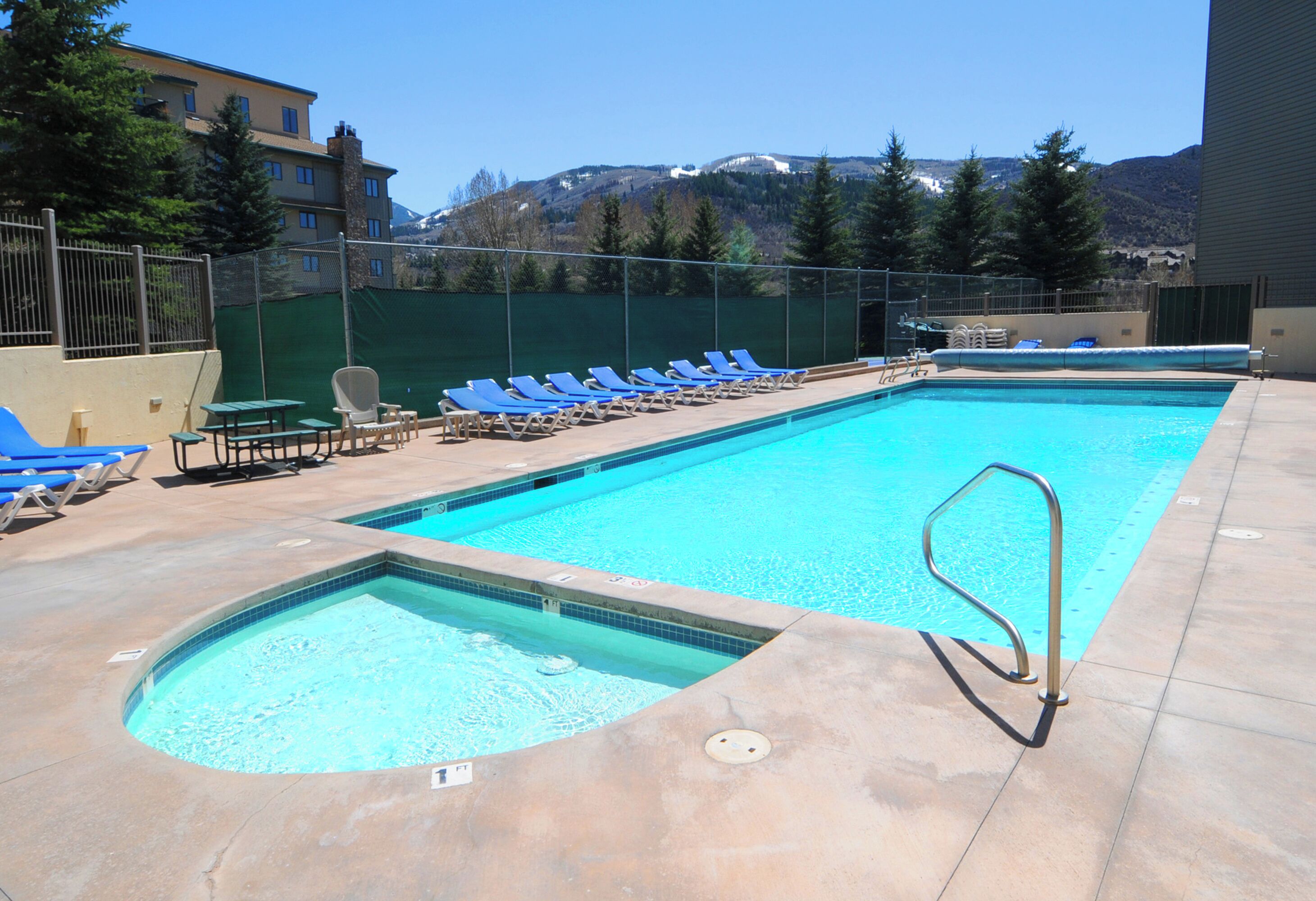 A rectangular swimming pool with a diving board, surrounded by blue lounge chairs and a green fence.