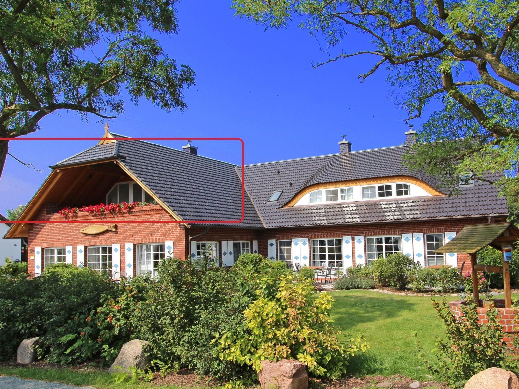 Ein zweistöckiges Haus mit einem geneigten Dach, einem Balkon mit roten Blumen und einem gepflegten Garten mit großen Steinen und Sträuchern.