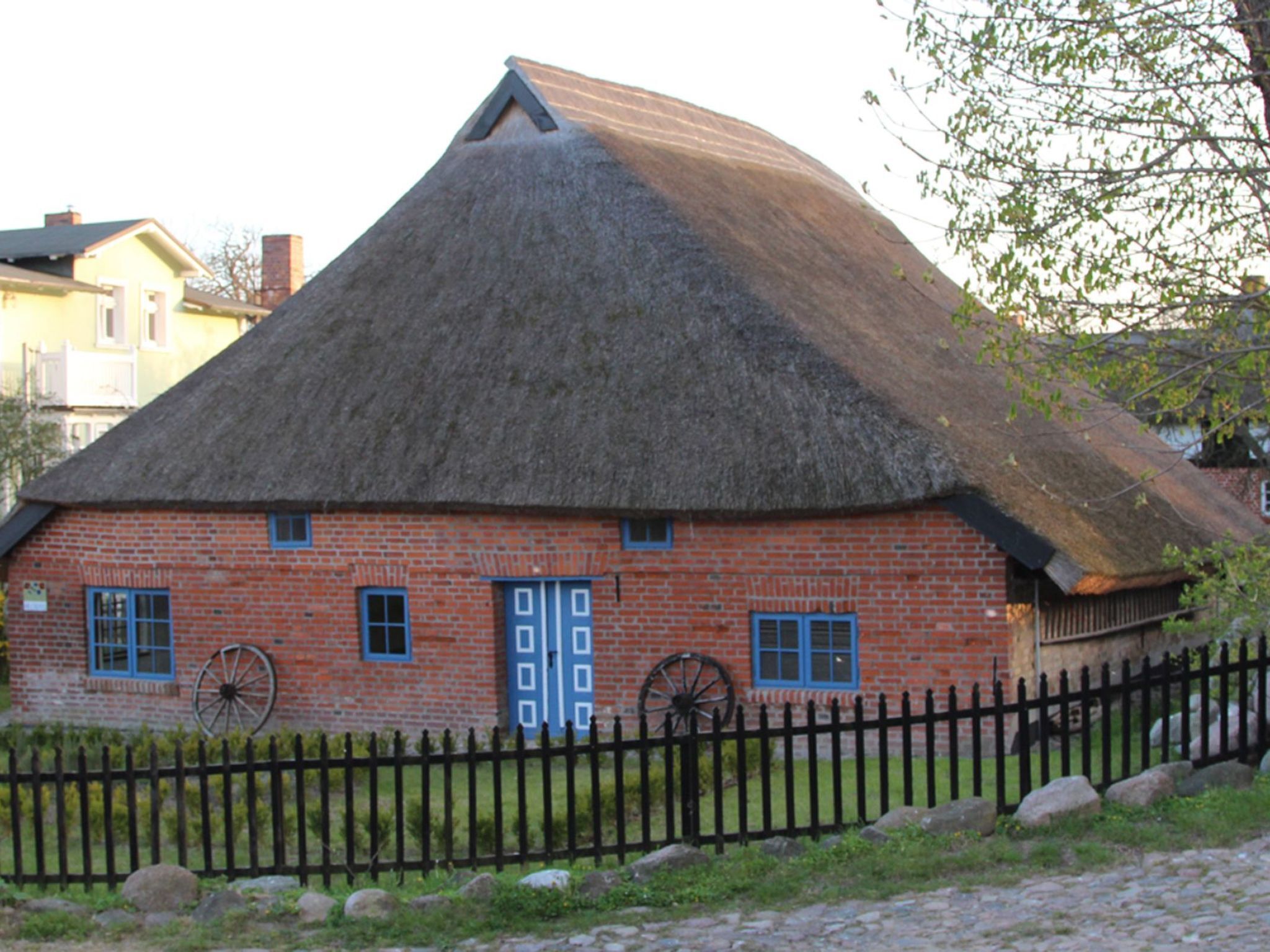 A traditional thatched-roof house with a brick exterior, a black metal fence, and a wheel in front.