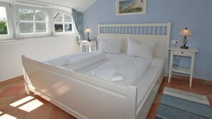 A bedroom with a white bed, two bedside tables with lamps, a window with curtains, and a painting on the wall. - Landhaus "To Hus" - Fewo Störtebeker mit Kamin by Interhome (Baabe)
