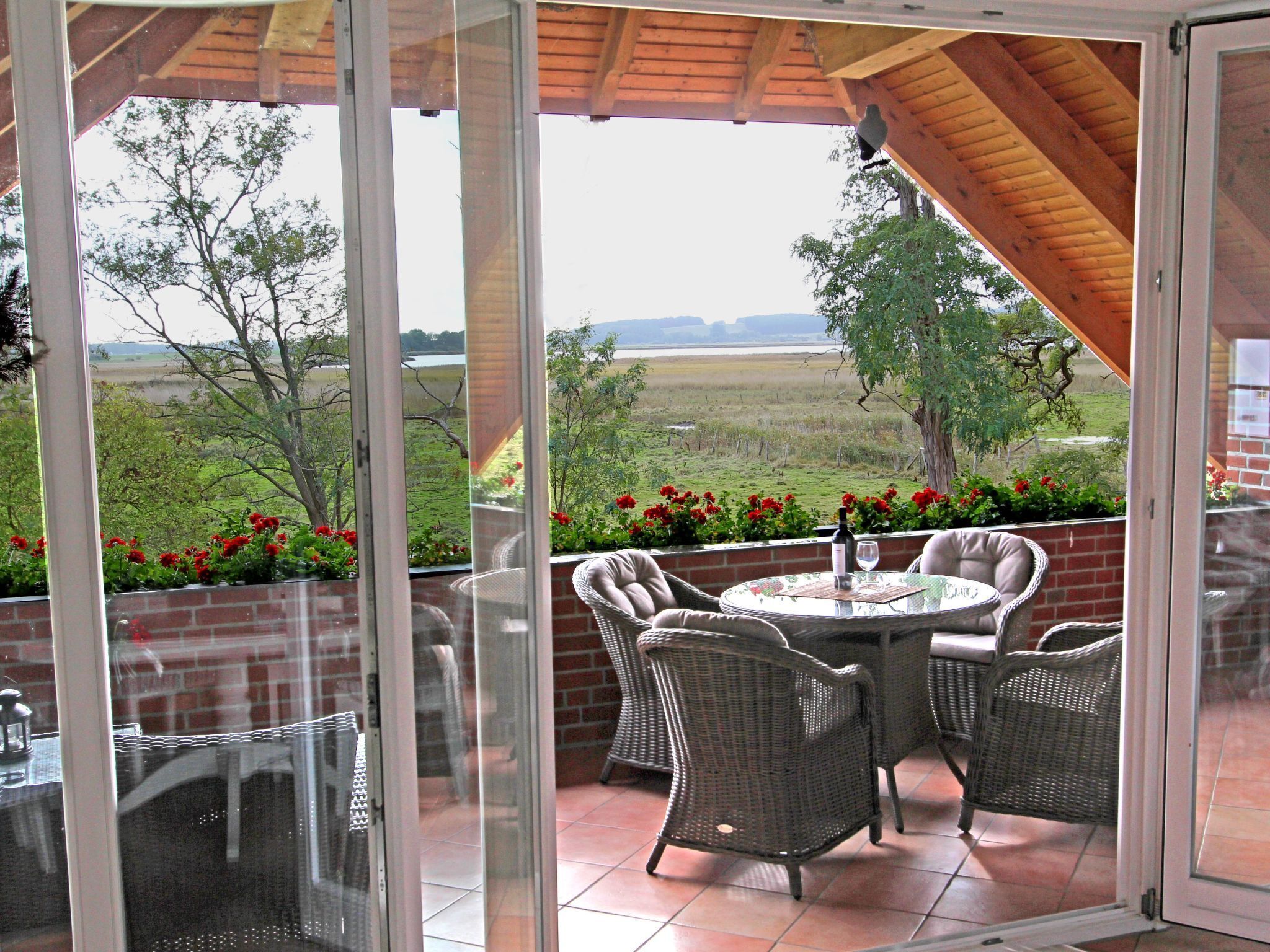 A covered patio with wicker furniture and a table, overlooking a grassy landscape with trees and red flowers.
