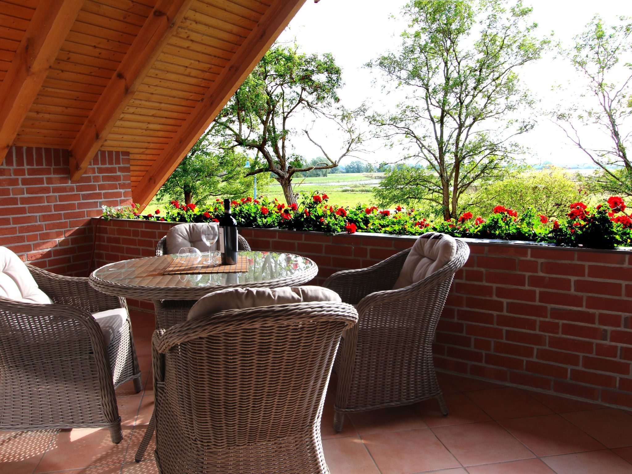 A covered patio with wicker furniture and a table, overlooking a garden with red flowers and trees.