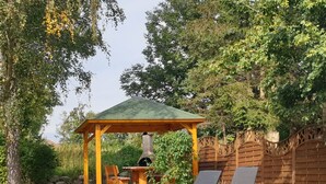 Un gazebo con tetto verde, circondato da una recinzione in legno e alberi. In primo piano, due poltrone da relax e un barbecue.