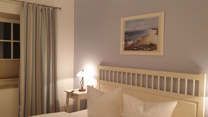 Un letto rifatto con lenzuola e cuscini bianchi, un comodino con una lampada, una finestra con tende e un quadro appeso al muro.