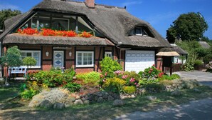 Ein traditionelles Haus mit Strohdach und weißer Garagentür, umgeben von einem gepflegten Garten mit verschiedenen Pflanzen und Blumen.