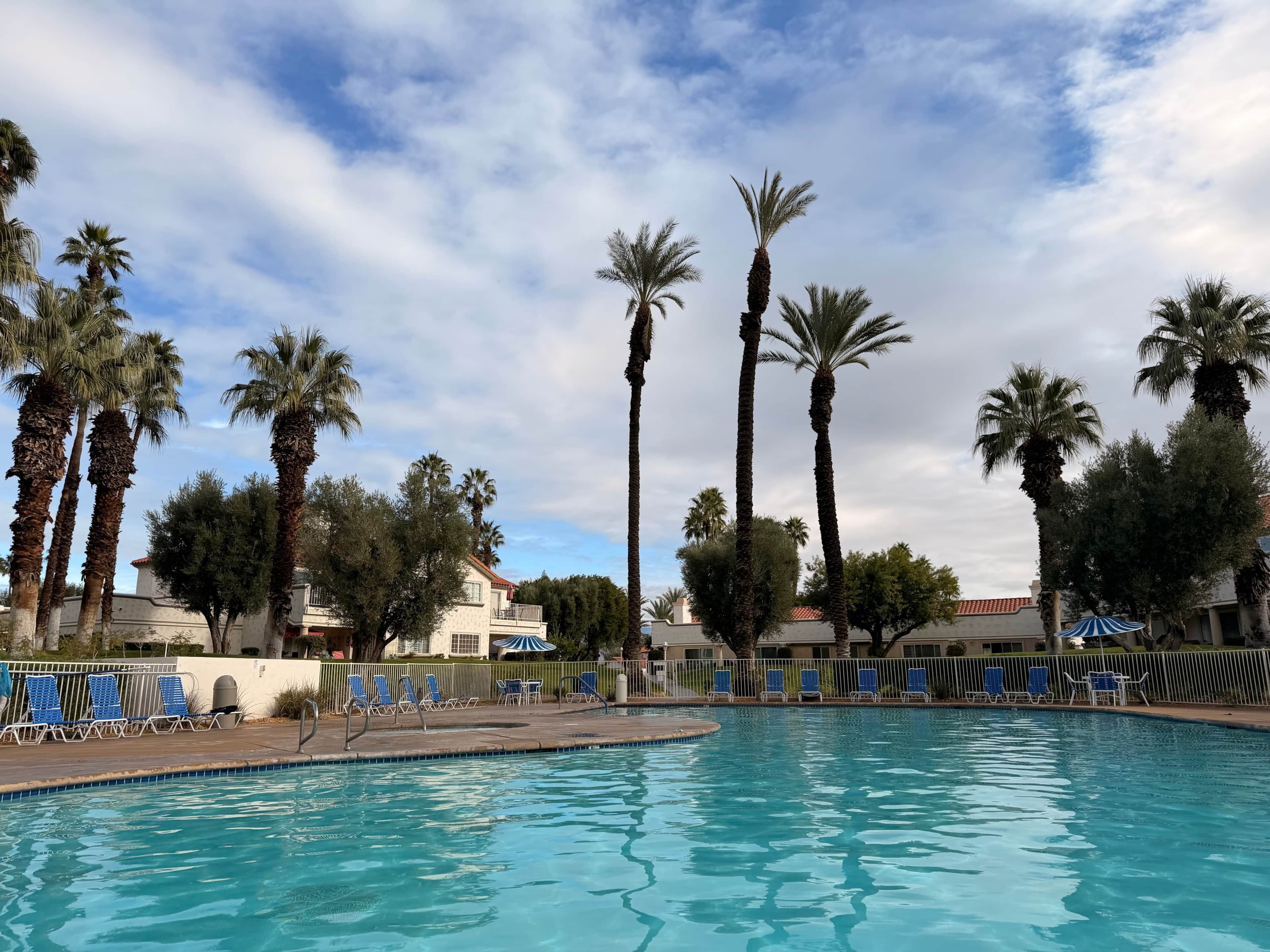 Una piscina con sillas reclinables azules, palmeras y un edificio con techo rojo al fondo.
