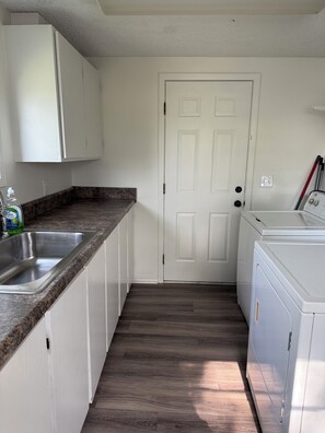 Una cocina con encimera de granito, lavaplatos de acero inoxidable, armarios blancos, una puerta blanca y una lavadora blanca.