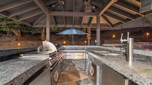 Une cuisine extérieure équipée d’un barbecue, d’un évier et d’appareils électroménagers en acier inoxydable, située sous une zone abritée dotée d’un sol en pierre.