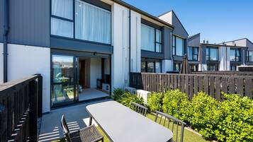 Een modern woongebouw met twee verdiepingen, voorzien van een patio met een tafel en stoelen, omgeven door groen en een houten hek.