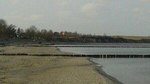 Il s'agit d'une zone côtière avec une plage de sable et une étendue d'eau calme. On y trouve des structures en bois le long du rivage, qui pourraient être des jetées ou des quais. Au fond, on aperçoit des bâtiments et des arbres, ce qui laisse supposer qu'il s'agit d'une zone résidentielle ou de loisirs.