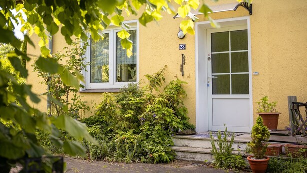 Un bâtiment jaune avec une porte et des fenêtres blanches, entouré de verdure et de plantes en pot.