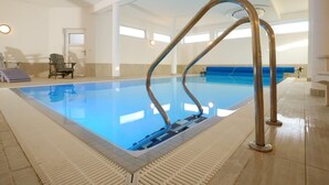 Una amplia piscina interior con superficie de agua azul cristalina, rodeada por un solárium con baldosas y una pared blanca.
