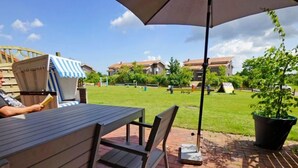 Eine Terrasse im Freien mit Tisch und Stühlen, einem Streifenmarkisenvorzelt und Blick auf eine Rasenfläche mit Gebäuden im Hintergrund.