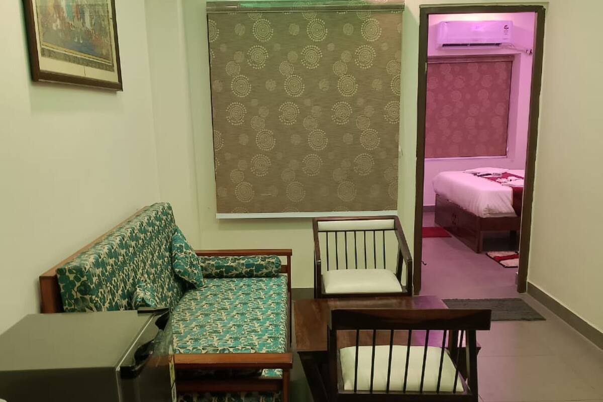 Ein kompaktes Wohnzimmer mit einer Couch, einem kleinen Tisch und einem Sessel. Im angrenzenden Zimmer befindet sich ein Bett, dessen Wand in Rosa gestrichen ist und eine 'Love'-Dekoration aufweist.
