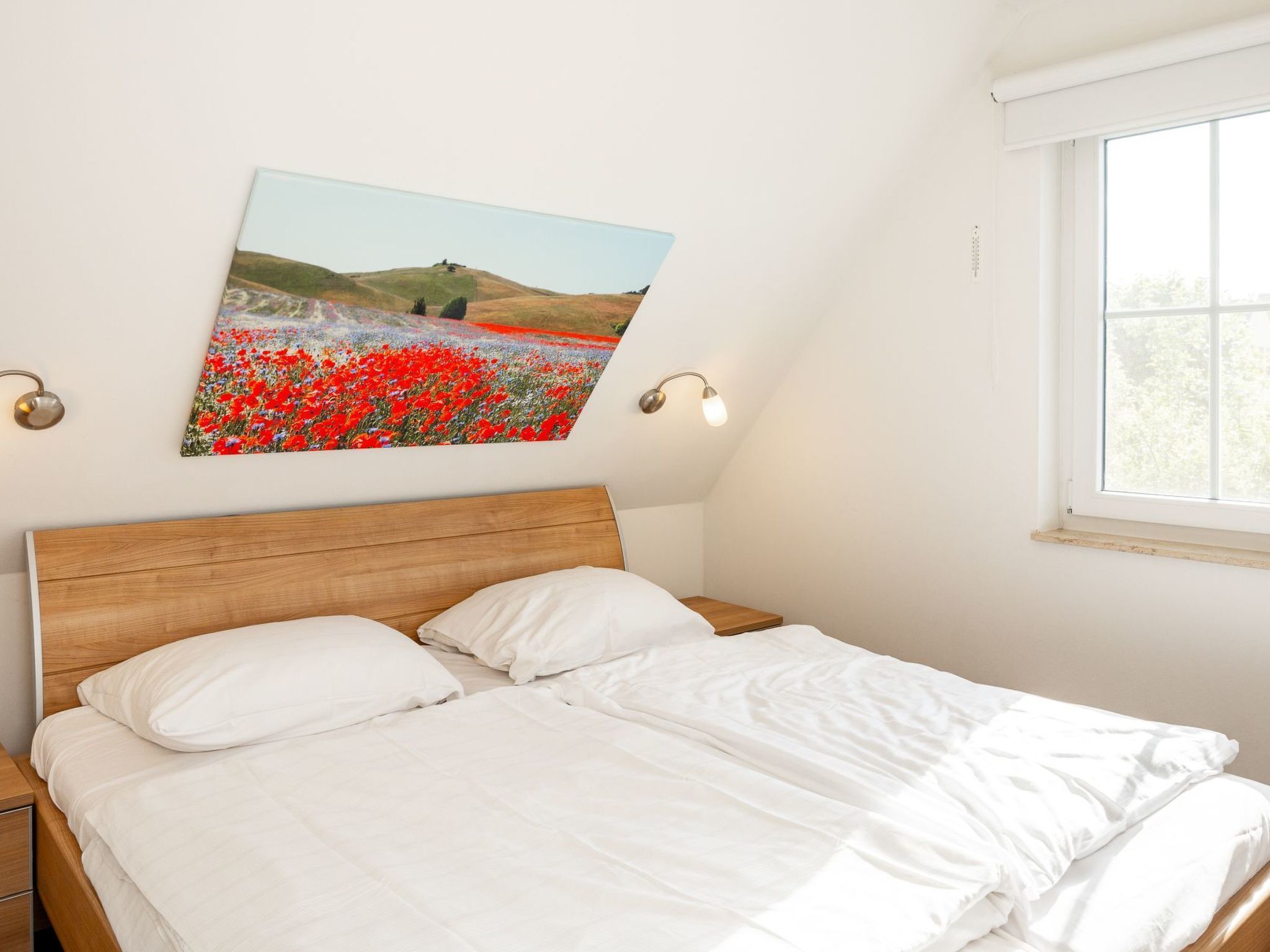 Een slaapkamer met een houten hoofdbord, wit beddengoed, een schilderij van een veld met rode bloemen en een raam met uitzicht op bomen.