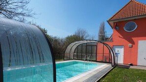 Ein Schwimmbecken mit Glasüberdachung, ein rotes Gebäude mit einer weißen Tür und ein klarer, blauer Himmel.