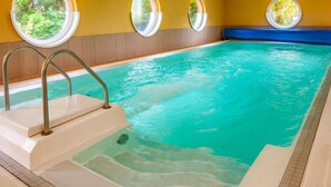 An indoor swimming pool with clear water, surrounded by yellow walls and windows with views of greenery.
